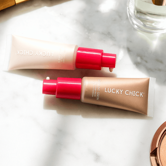 Lucky Chick Dawn Glow and Radiant Glow Fruit-Infused Illuminizers with pink caps on a marble surface in natural light.